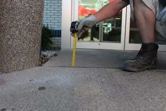Person wearing gloves and boots measures the height difference between two concrete sidewalk slabs using a yellow measuring tape outside a building entrance.