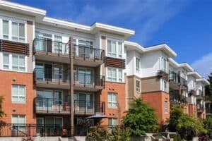 Modern, multi-story apartment building with large windows, balconies, and brick and white panel exterior, set against a blue sky with green landscaping in front.