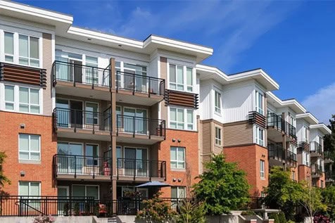 Modern, multi-story apartment building with large windows, balconies, and brick and white panel exterior, set against a blue sky with green landscaping in front.