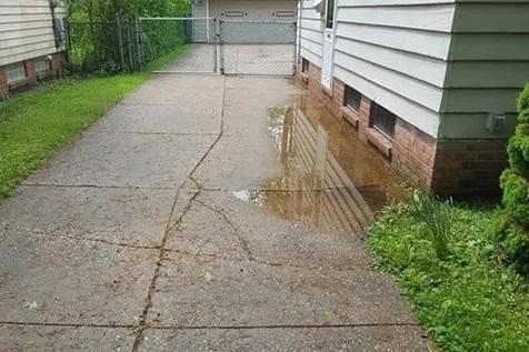 A puddle of water is collected along the edge of a cracked concrete driveway beside a house, near a metal gate and some grass.