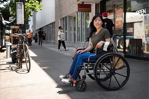 A woman in a wheelchair is on a city sidewalk near storefronts and bicycles. Other pedestrians walk by as she looks slightly to the side.