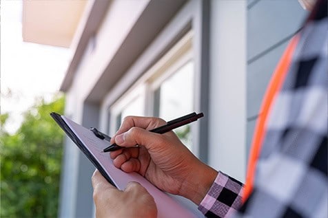 Person holding a clipboard and writing with a pen outside a building, wearing a checkered shirt and orange safety vest.