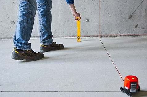 A person in jeans and sneakers uses a bubble level to check alignment on a floor marked with a red laser line, next to a laser level device.