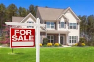 A two-story house with a “For Sale” sign in the front yard, surrounded by grass, bushes, and trees in the background.