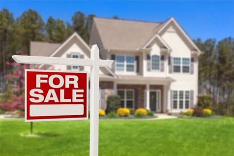 A two-story house with a “For Sale” sign in the front yard, surrounded by grass, bushes, and trees in the background.