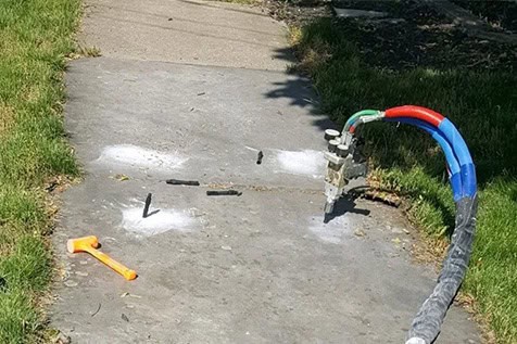 A robot arm with colored cables is drilling into a concrete sidewalk; white powder, debris, and an orange tool are scattered nearby.
