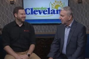 Two men are seated indoors, talking in front of a TV displaying the “New Day Cleveland” logo.