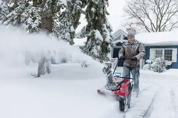 A person uses a snow blower to clear a driveway in front of a house surrounded by snow-covered trees and yard.