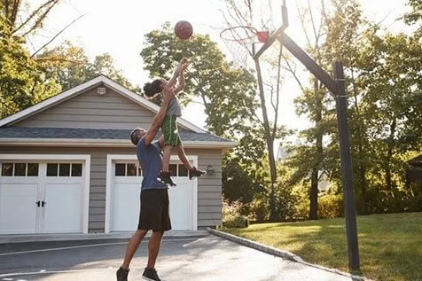 An adult lifts a child to help them shoot a basketball into a hoop on a driveway in front of a house.