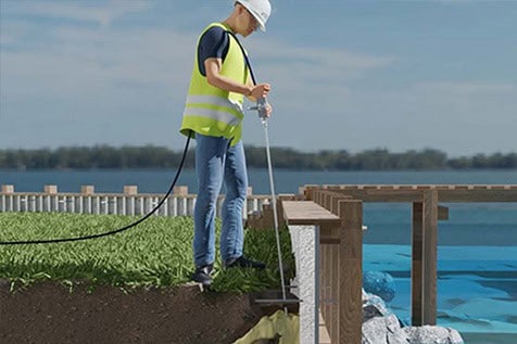 A worker in a safety vest and helmet uses equipment to inspect or repair a dock near the water, standing on grass by the edge.