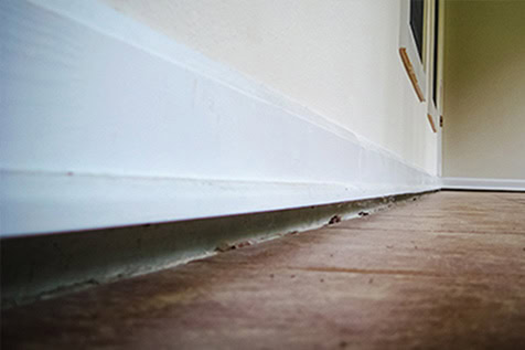 Close-up view of a white baseboard along a wall with visible gaps and debris between the baseboard and brown floor.
