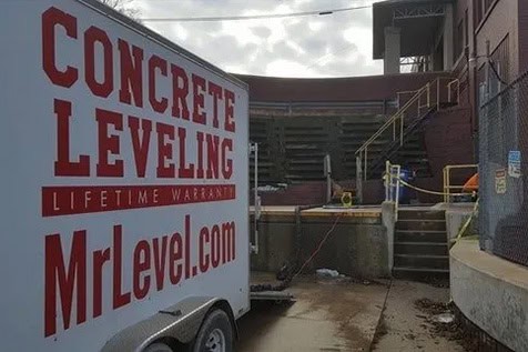 A trailer labeled “Concrete Leveling MrLevel.com” is parked near concrete steps and a building under repair on a cloudy day.