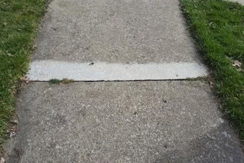 A concrete sidewalk with a single light-colored rectangular patch contrasting with the surrounding darker concrete; grass borders both sides.