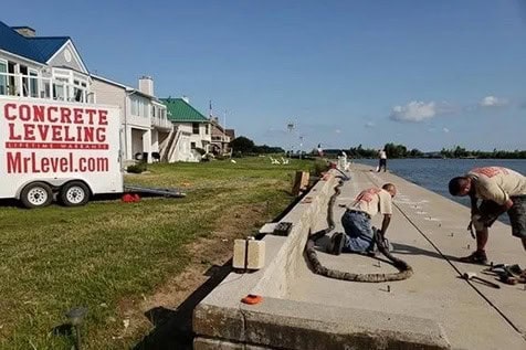 Two workers repair a concrete walkway beside a canal, with a trailer labeled “CONCRETE LEVELING MrLevel.com” parked on the grass nearby.