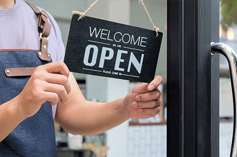 A person in an apron hangs a black sign that says “Welcome We Are Open” on a glass door, indicating that the business is open.