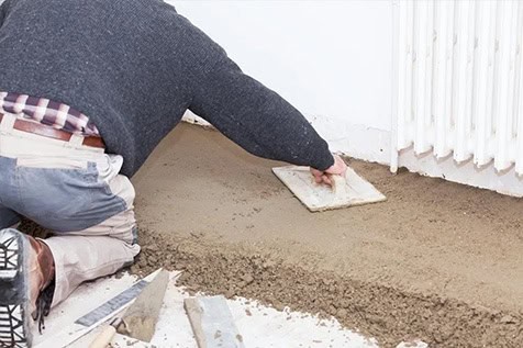 A person kneels on the floor, spreading fresh concrete with a trowel next to a wall and radiator. Tools are visible nearby.