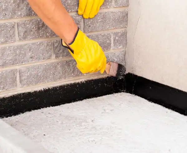 A person wearing yellow gloves applies black sealant with a brush to the joint between a brick wall and a concrete floor, an important step in basement waterproofing.