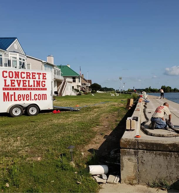 A worker repairs a concrete seawall next to a grassy area and houses, with a trailer labeled “Concrete Leveling MrLevel.com” parked nearby.